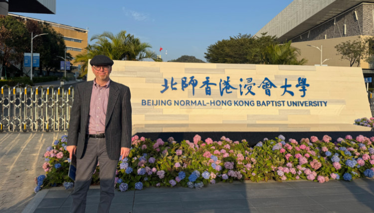 Gonzaga University philosophy professor visiting Beijing Normal–Hong Kong Baptist University campus in Zhuhai, China. The image shows the university’s entrance sign with bold blue Chinese and English lettering, surrounded by colorful flowers and modern academic buildings under a clear blue sky.