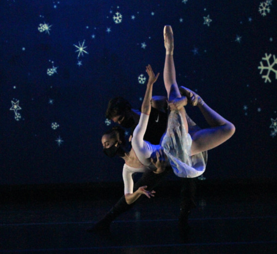 Two masked dancers, one holding the other up while she poses with legs pointing to the ceiling. Background is blue with snowflakes.
