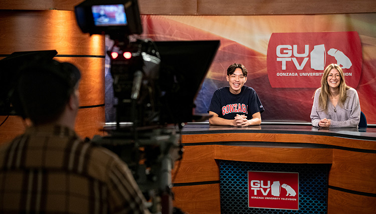Two student broadcasters at the broadcast desk with a camera operator in the background.