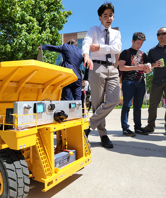 A young man points to a very small tractor on a sidewalk with a panel on its front