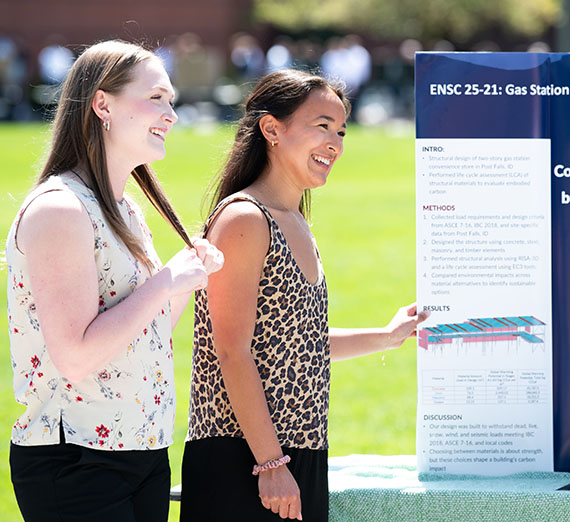 Two female students smile at a visitor to their academic poster outside on a sunny day