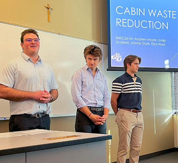 Three students stand by a projection screen that reads Cabin Waste Reduction