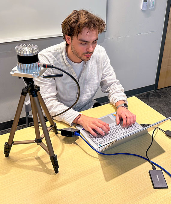 A young man types into a laptop connected to electronic components on the table and on a stand