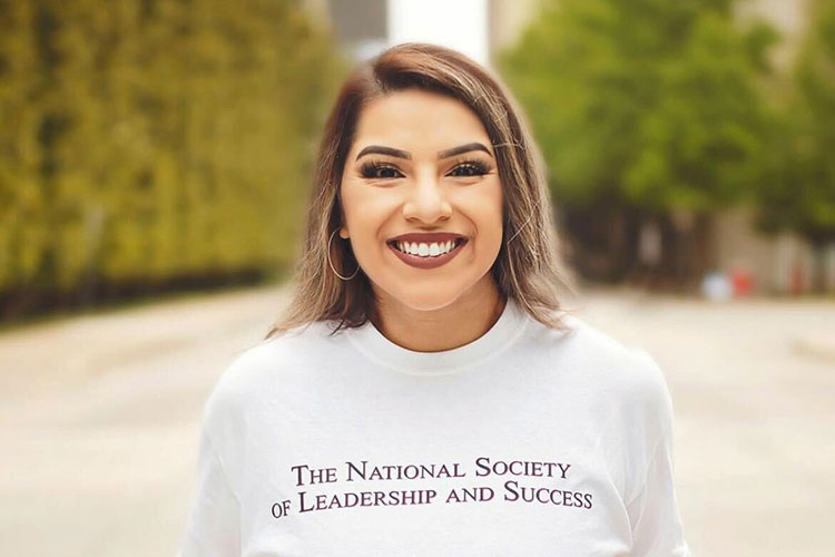 Smiling student standing outdoors wearing a white T-shirt with the words 'The National Society of Leadership and Success' printed on the front.