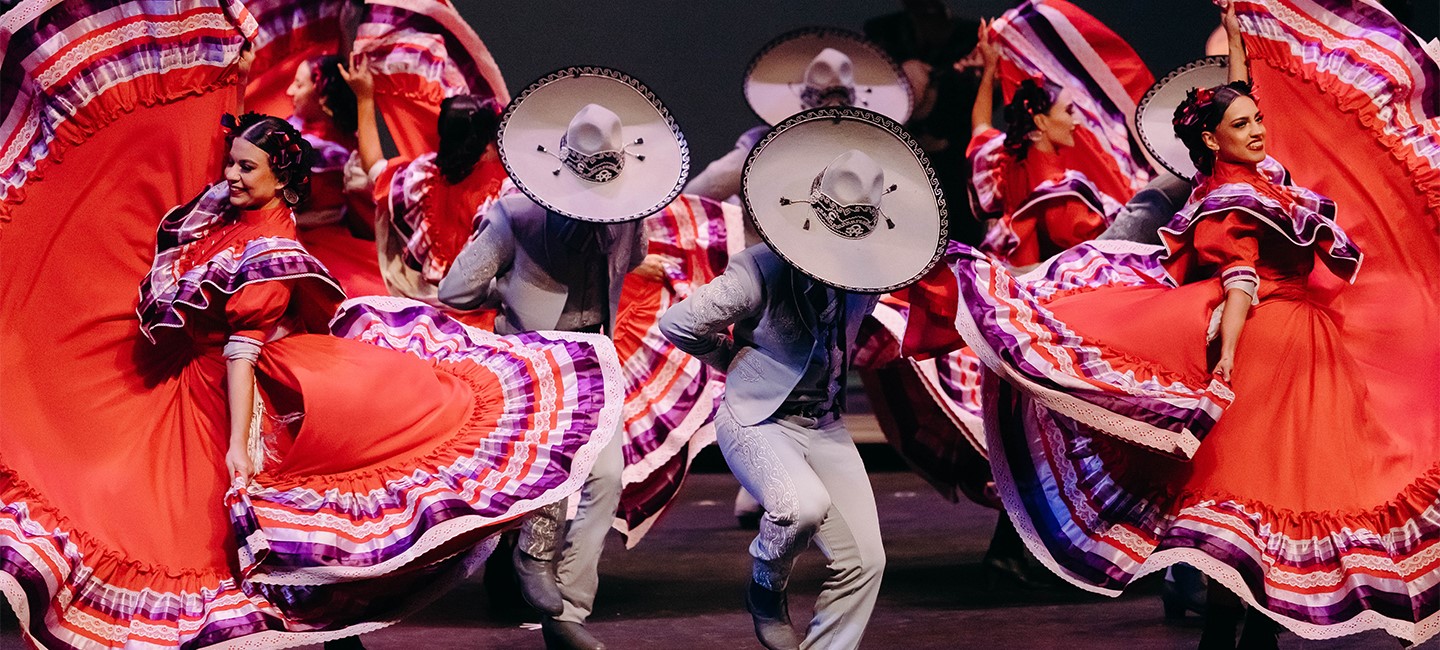 Performers on stage dancing in red dresses or suites with large brimmed hats. 