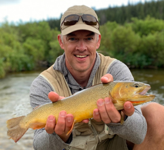 Daniel Vimont holds a trout with a river and trees in the background
