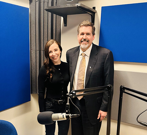  Sarah Schwering, Presidential Communications Specialist and Thayne McCulloh, President stand side by side in a podcast room with blue background and a microphone in the foreground.