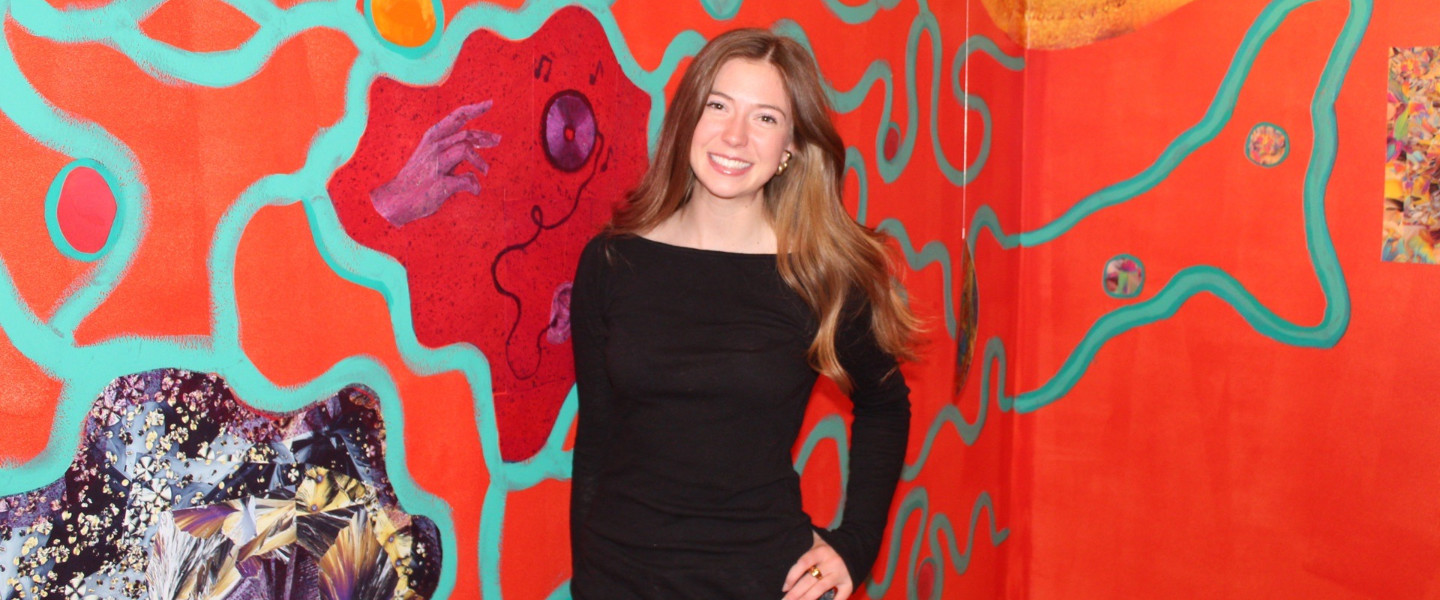 Gonzaga University Neuroscience and Art Student Ava Knap in front of her exhibit "Where's My Mind: Neuroscience Meets Art An Abstract Installation of the Mind" which is a bright orange wall with neurological structures painted on it. 