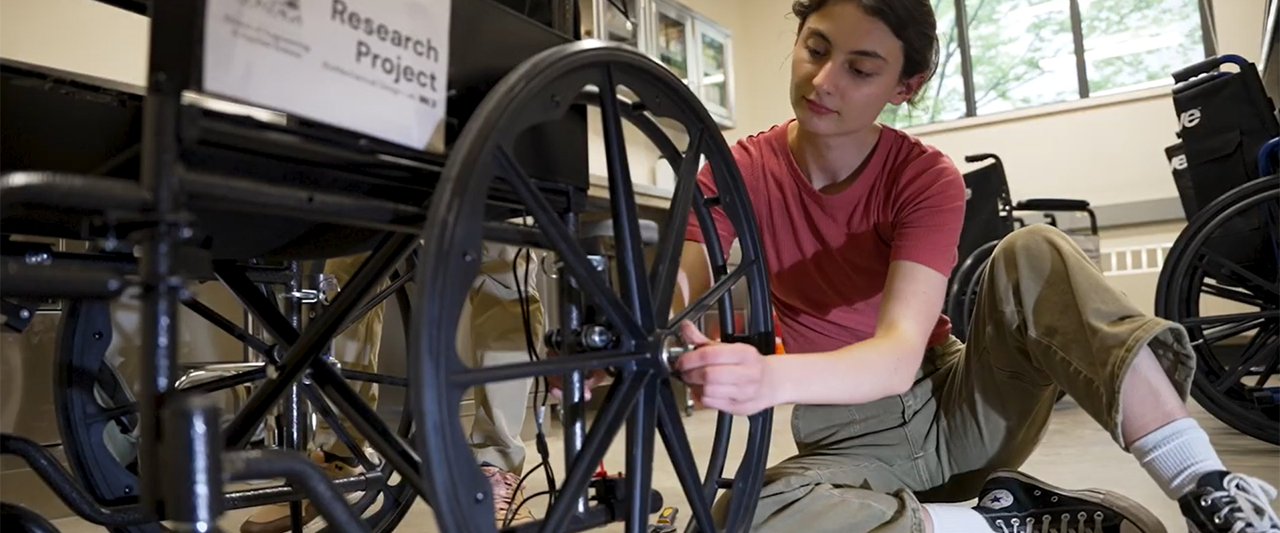 A person in a red shirt and olive pants kneels, working on a black wheelchair's wheel. A "Research Project" sign is visible on another wheelchair nearby.