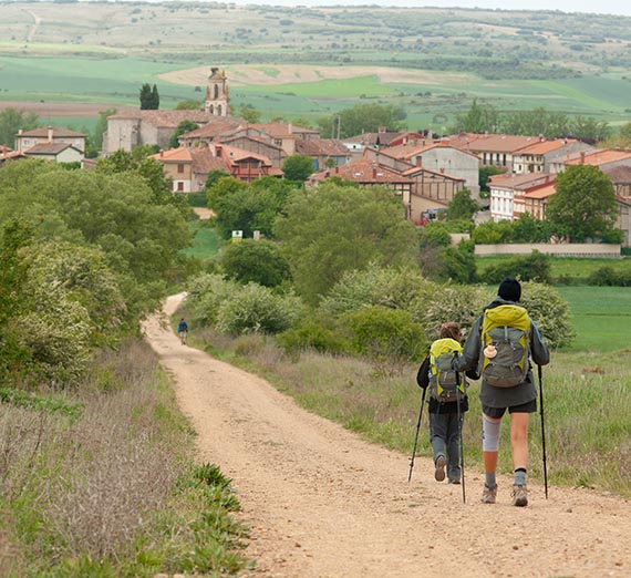 An adult and a child walk down a dirt road leading into town shown in the distance
