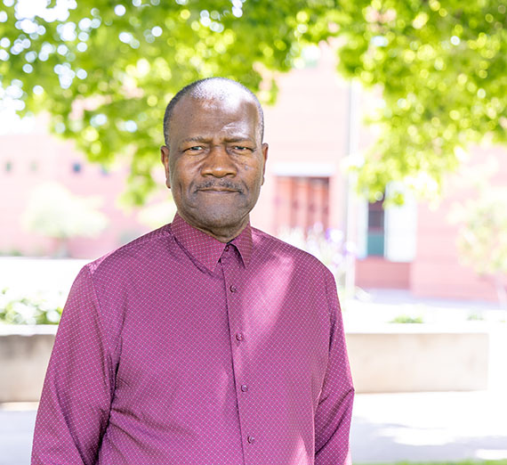 Male student wearing red button up standing with red brick building and trees in the background 