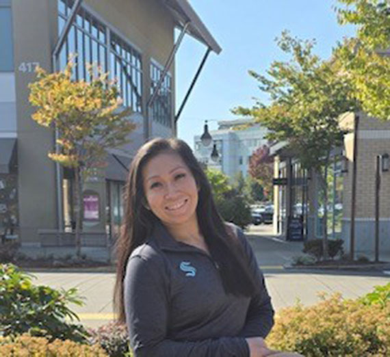 Female with long brown hair standing outside of a building with trees around it and wearing a Seattle Kraken hockey shirt. 