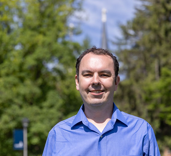 Male student wearing blue button up with trees and church spire in the background 