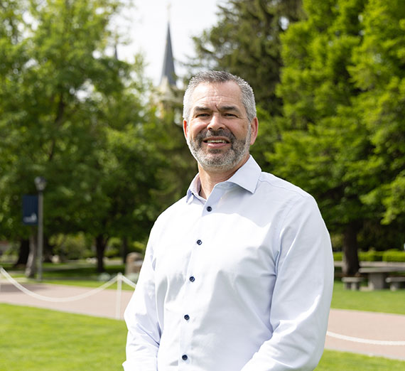 Male with a white button up standing in front of trees and college campus 