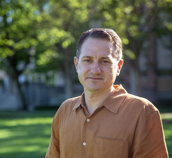Man in button up orange shirt with greenery of campus behind him 