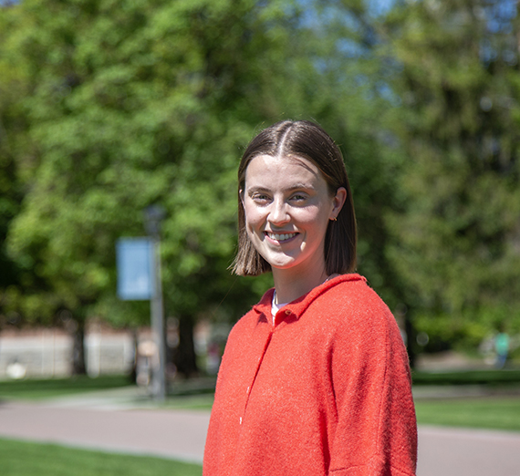 Female student in a red sweater standing on campus with trees in the background 