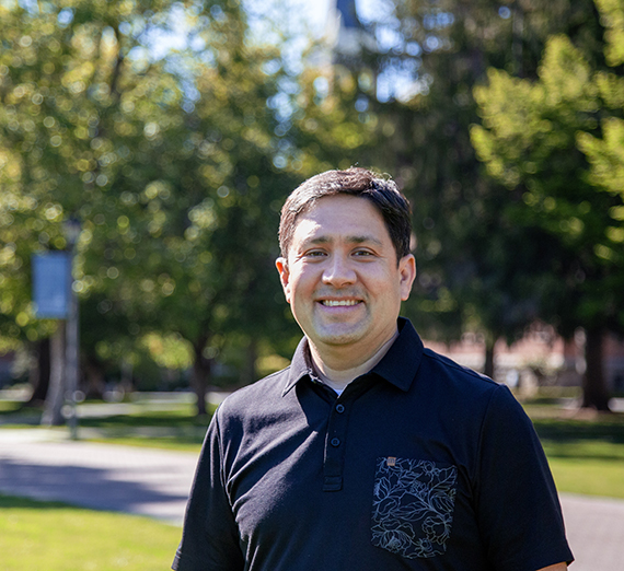 Male student wearing black polo stands with trees, brick walkway, and brick buildings in background 