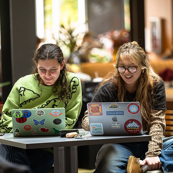 Two students sitting side-by-side with their laptop computers on a small table.