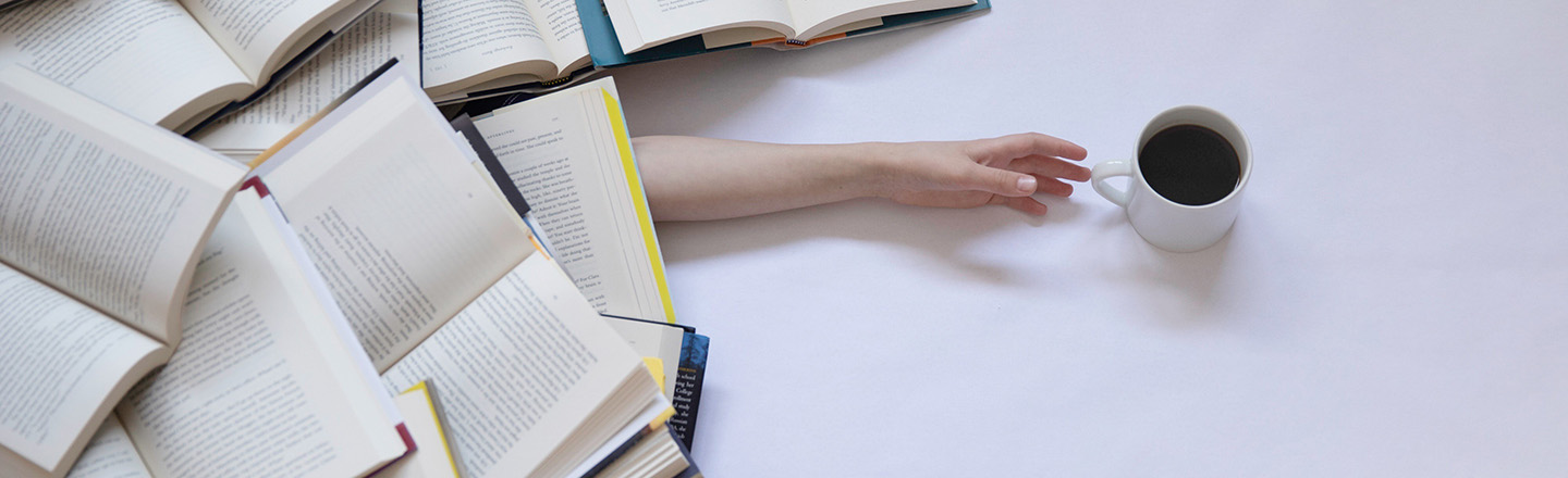 Person under a pile of books reaches for a cup of coffee.