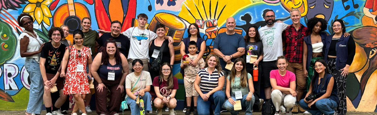 Group of 23 people standing in front of a colorful, painted wall.