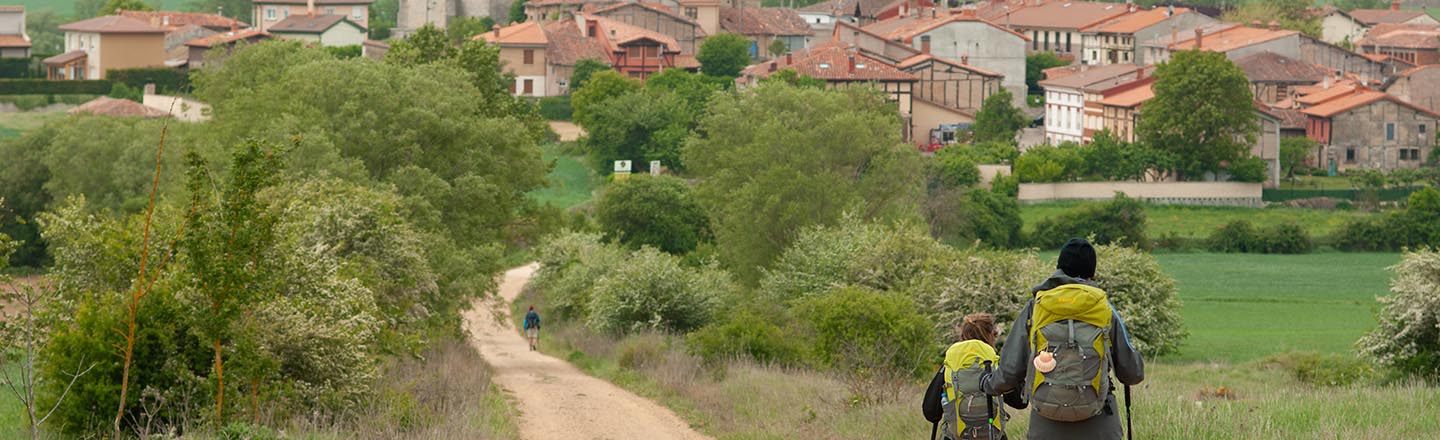 An adult and a child walk down a dirt road leading into town shown in the distance