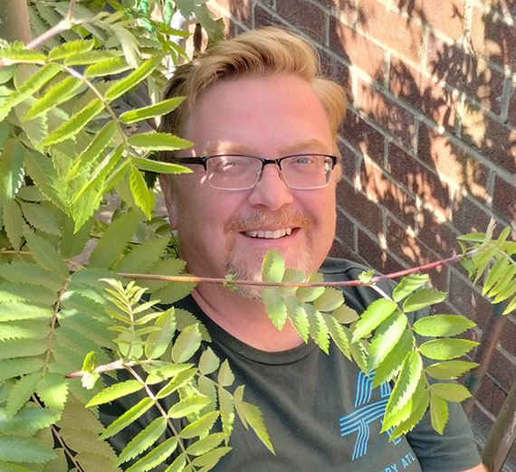 Josh Hogan wearing glasses beside a brick wall with vines in the foreground of the photo.