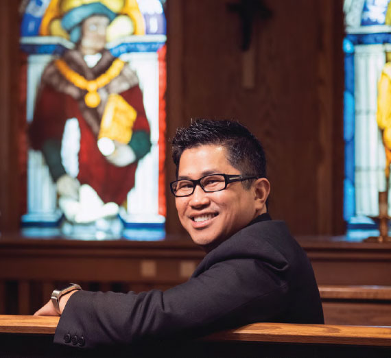 A man smiles while seated in a church, with stained glass windows behind.
