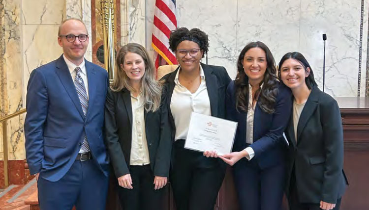 A group of five individuals proudly posing in a grand hall, with one holding a certificate, next to an American flag.