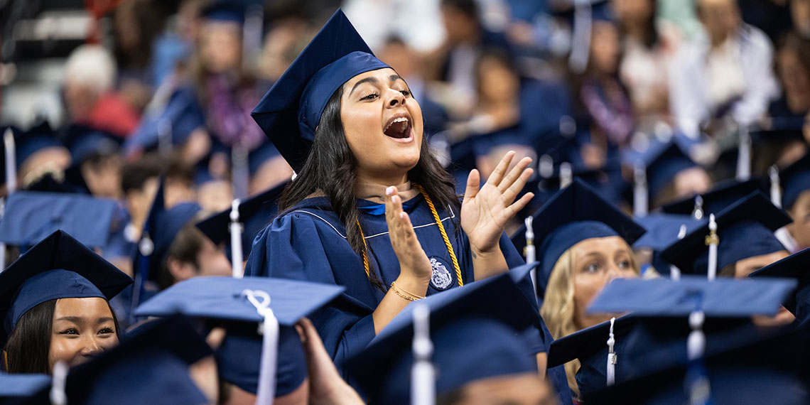 A female student stands among sitting fellow graduates at commencement ceremony