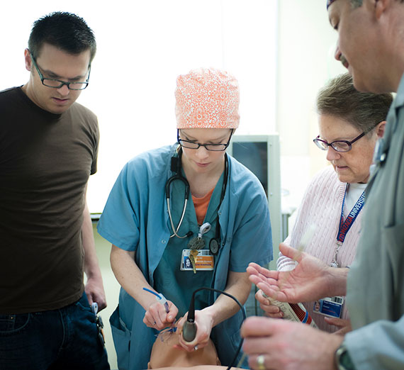 A woman in medical scrubs places a tube down a medical training dummy's throat while students watch