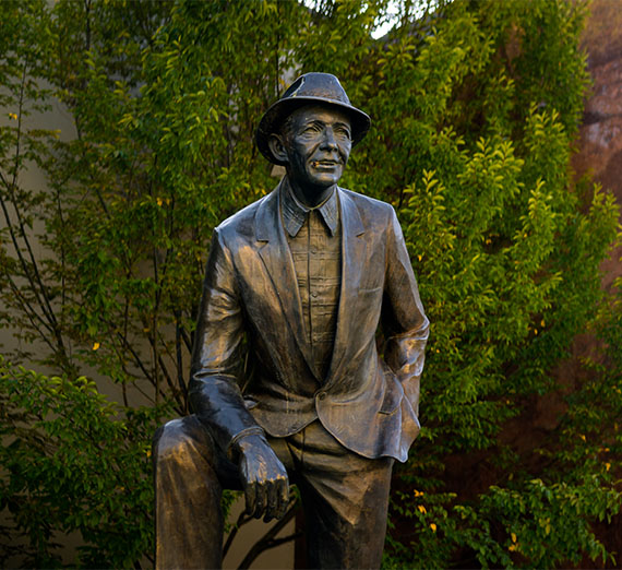 Statue of Bing Crosby in a suit and hat, smiling with one hand on his knee, surrounded by trees.