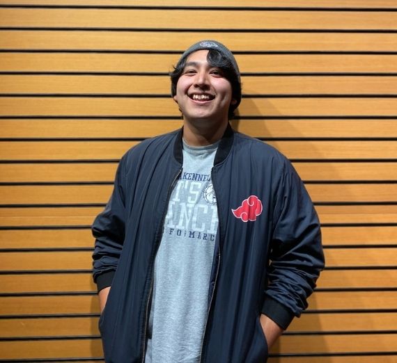 Current student standing against a wood-paneled wall. He is wearing a gray beanie, a navy windbreaker, and a gray Kennel Club t-shirt. He is smiling.