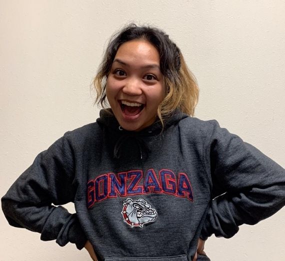 A photo of a current Gonzaga student smiling. She is wearing a gray sweatshirt that spells GONZAGA in navy letters outlined by red. There is a bulldog also on the sweatshirt.
