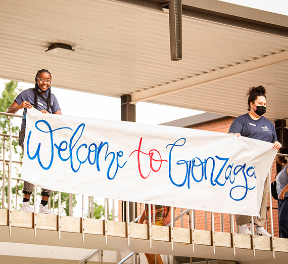 Two student leaders hanging a "Welcome to Gonzaga" banner outside their residence hall as students move in during 2021 orientation and move-in.