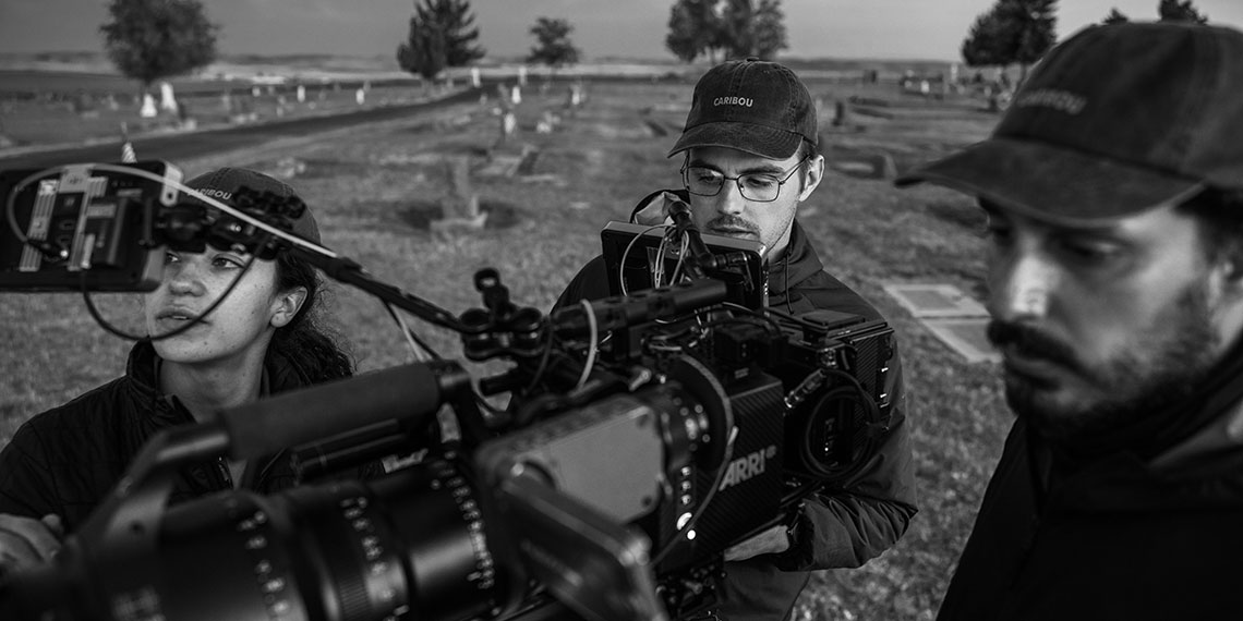 A woman and two men stand behind a film camera in a cemetary