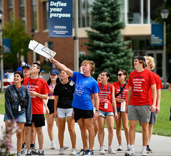 A student orientation leader points out a building to new students standing on the sidewalk