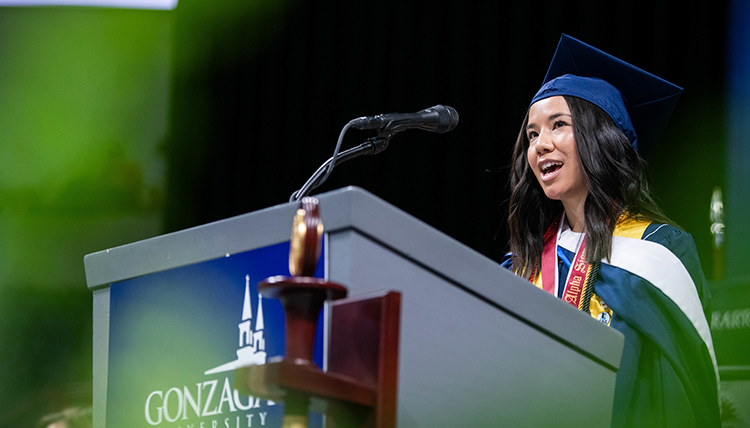 A young woman speaks at a podium while wearing a graduation cap