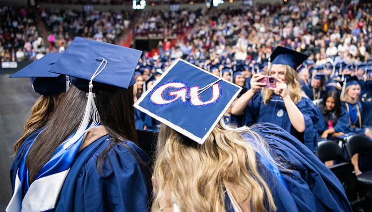 A woman in commencement regalia takes a picture of two friends