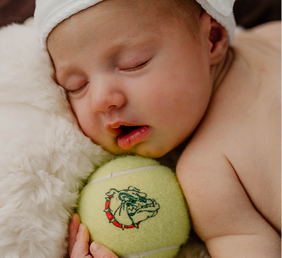 Baby asleep with a tennis ball with a Zag logo on it.