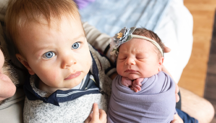 A blue-eyed little boy holds his newborn baby sibling.