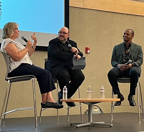 A woman, a priest and a man sit on a stage holding microphones