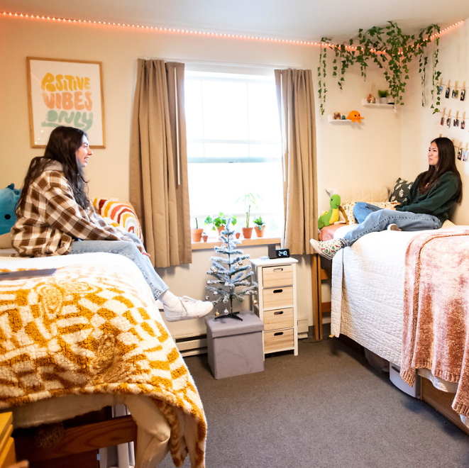 Two female college students sitting in their dorm room chatting. They are both smiling and are both sitting across from each other on their raised beds. On the left, the student has dork brown hair and sits on her bed wearing a yellow and brown flannel shirt and blue jeans. Her bedding is white with a yellow blanket. The student on the right also has dark black hair and is wearing a green sweater and blue jeans. Her bed has white bedding and a pink blanket. A plant hangs above the head of the bed on the left. In between their two beds, is a white nightstand and a grey ottoman with a mini Christmas tree on top. 