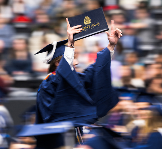 A man in commencement regalia holds up his degree with Gonzaga University written on it