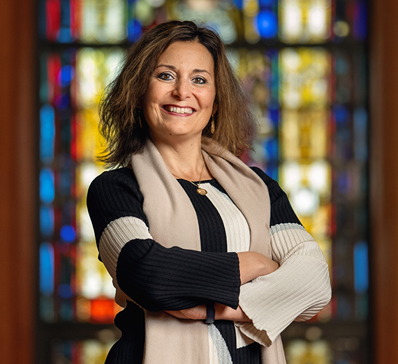 Dr. Passerini poses with her arms crossed in front of a stained glass window. 