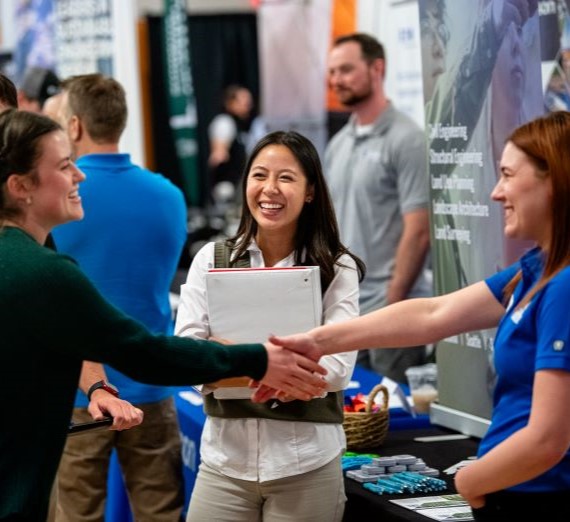 A Gonzaga student in a green sweater shaking hands with an employer at a career fair wearing a blue polo with another employer smiling watching the interaction.