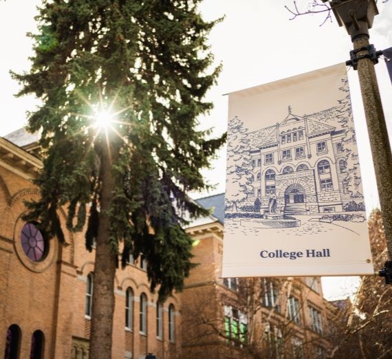 A sign for College Hall on a light post with a drawing of College Hall in blue also on the sign. In the background is a pine tree with the sun shining through it and the northeast side of College Hall.