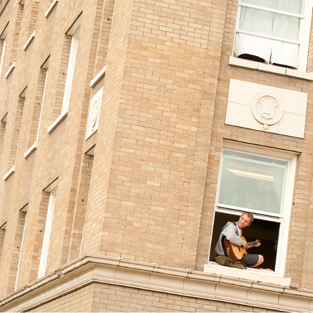 Student sitting in a window sill of DeSmet Hall on Gonzaga's campus. The student is wearing a grey tee shirt and grey shorts. He is playing the guitar and smiling. The building around him has a tan brick with white stone accents.