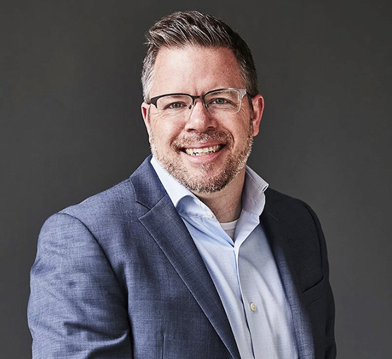 Alumni, Ray Harter. White male wearing button up shirt with blazer and glasses with a grey background.