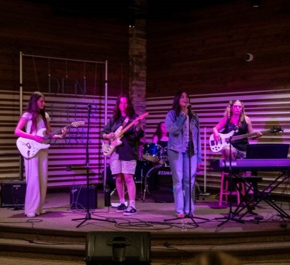 A group of students playing in a band in the Hemmingson Den for the Wednesdays' "Den After Dark" performance series. The band has four female students playing various instruments including the drums, two electric guitars, and one bass guitar,, as well as a singer wearing a denim jacket.