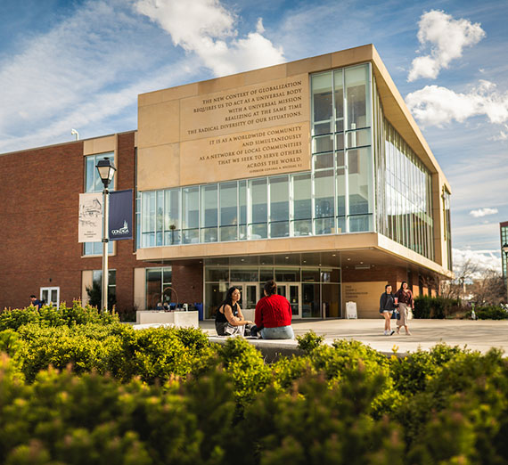 Photo of brick and glass building with bushes in the front and students walking around